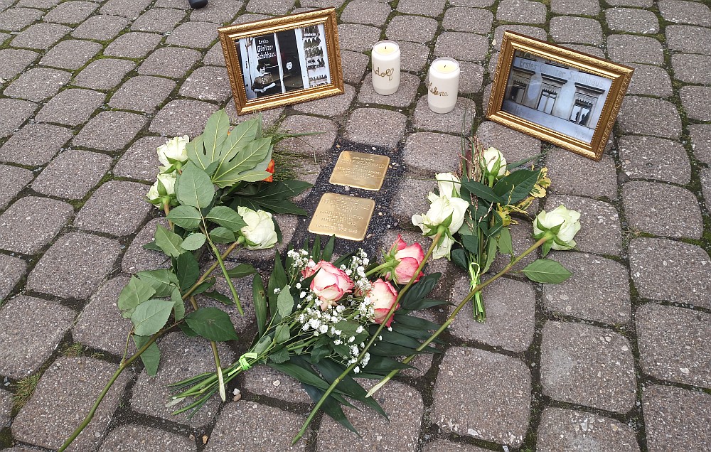 zwei Kerzen und Blumen schmücken die neuen Stolpersteine, im Hintergrund Bilder des Geschäfts in den 30er Jahren